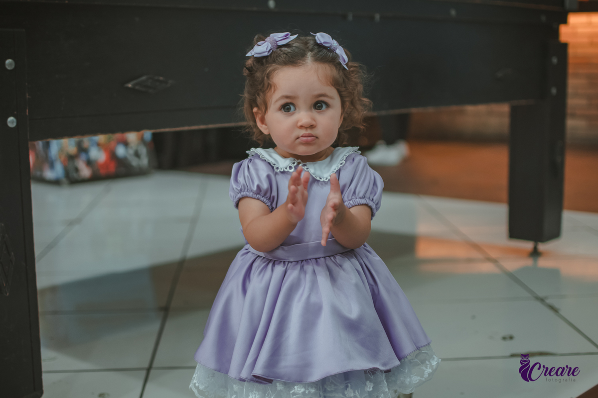 Uma bebê usando vestido roxo e batendo palmas durante seu aniversário de 1 ano.