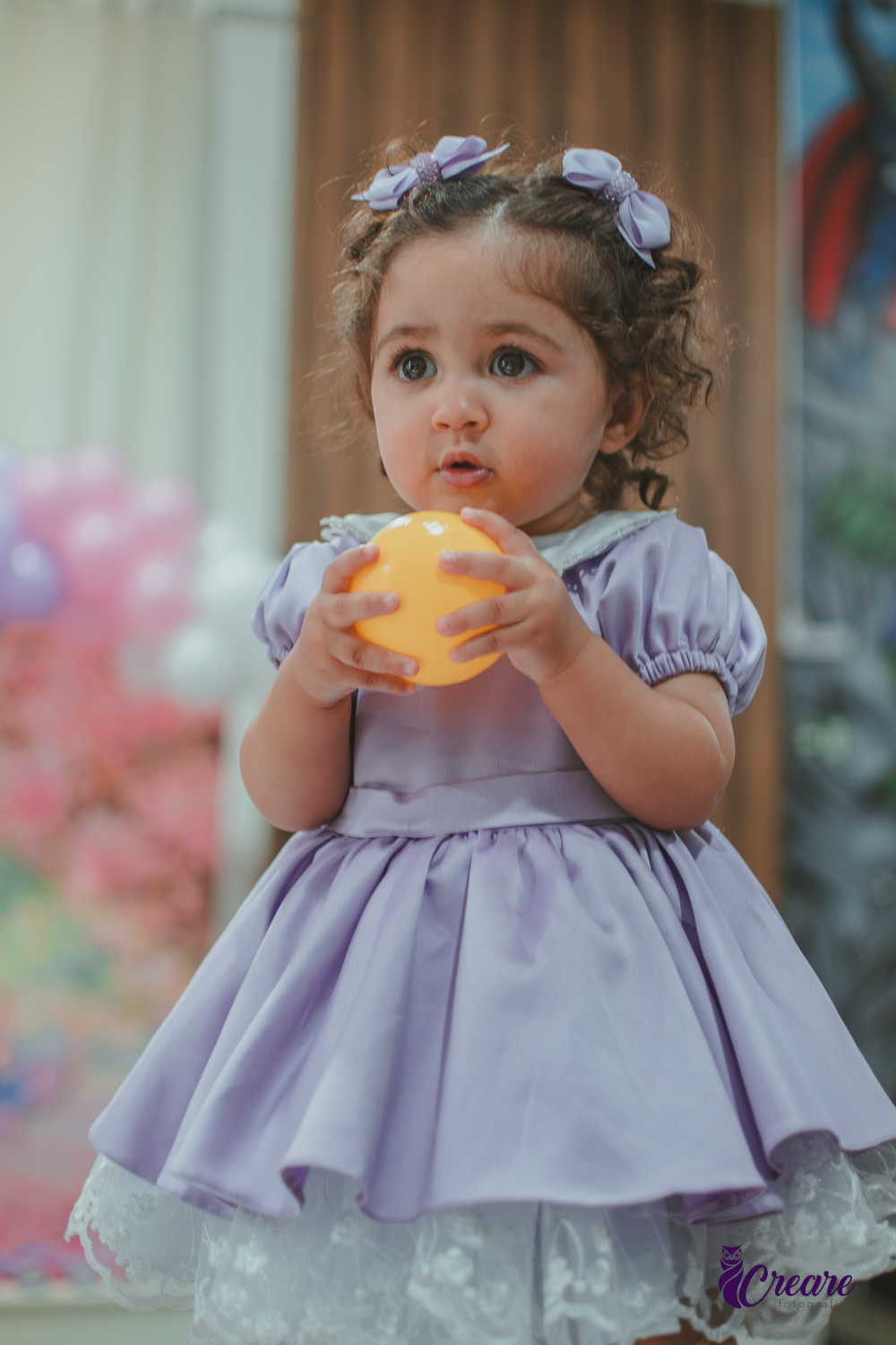 Uma bebê usando vestido roxo e segurando uma bolinha amarela, durante seu aniversário de 1 ano.
