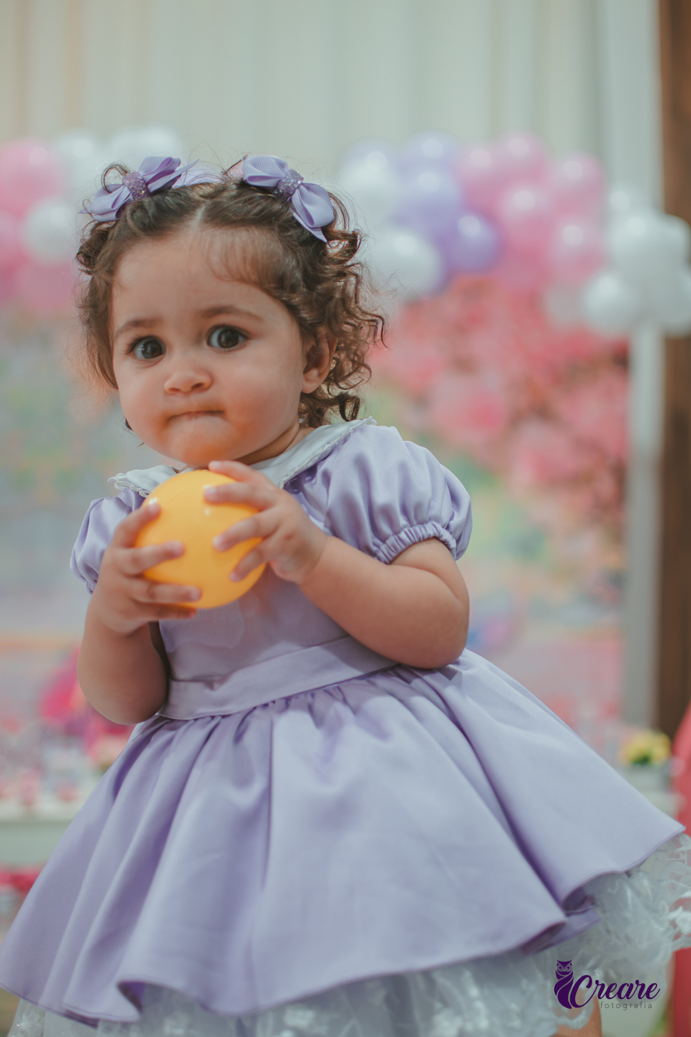 Uma bebê usando vestido roxo e segurando uma bolinha amarela, durante seu aniversário de 1 ano.