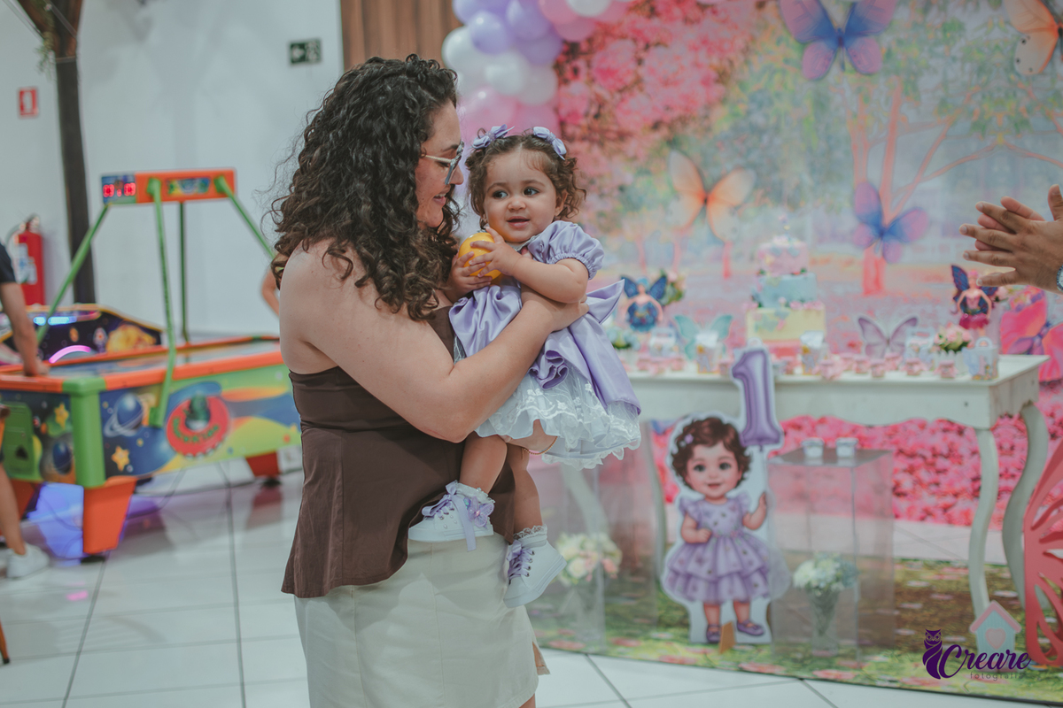 Uma mulher segurando sua bebê, no salão do buffet, durante festa de aniversário de 1 ano.