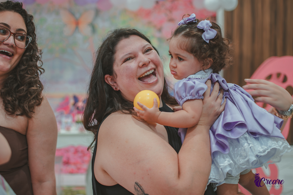 Uma mulher segurando sua bebê e sorrindo, no salão do buffet, durante festa de aniversário de 1 ano.
