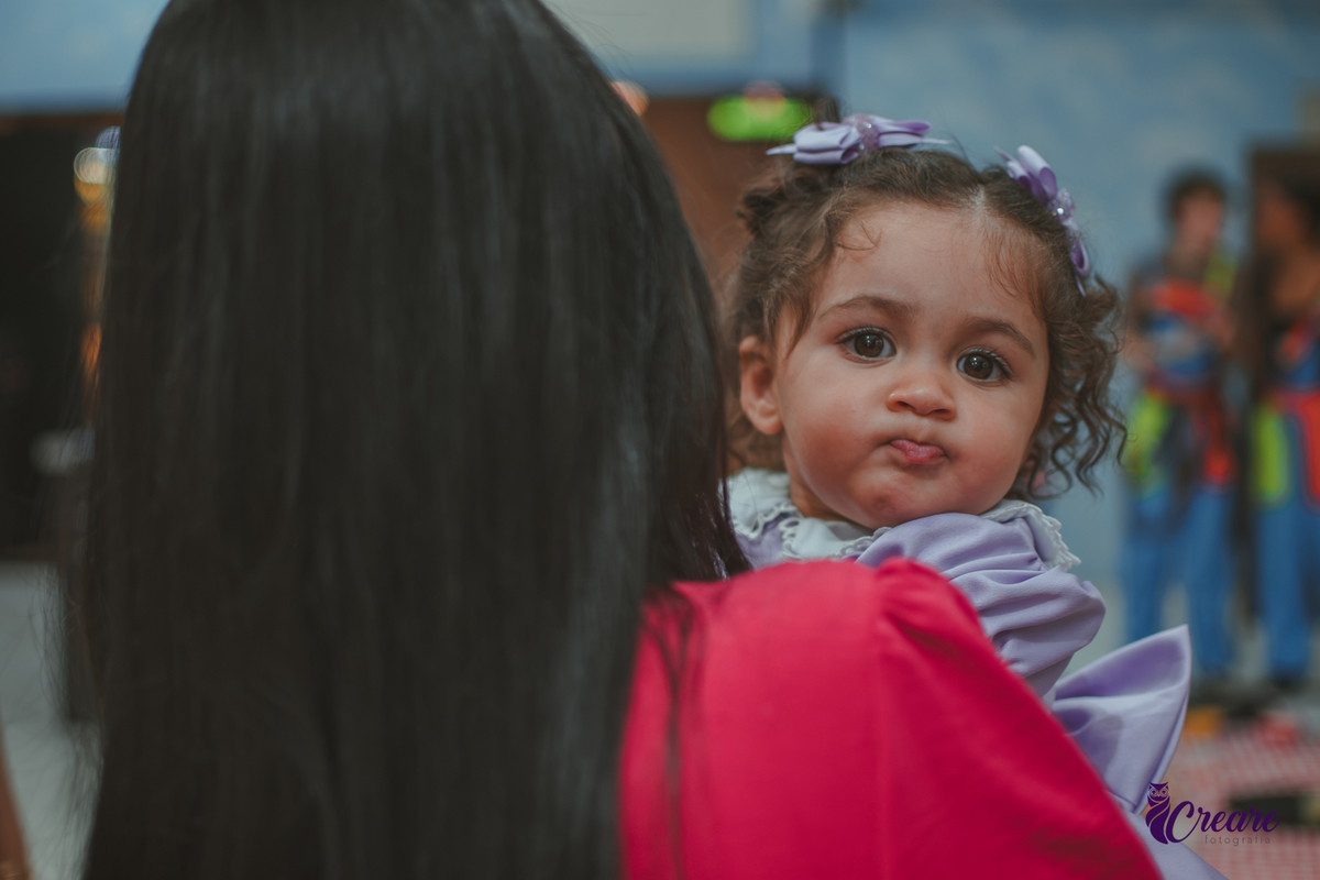 Uma bebê olhando por cima do ombro de uma mulher, durante festa de aniversário de 1 ano.