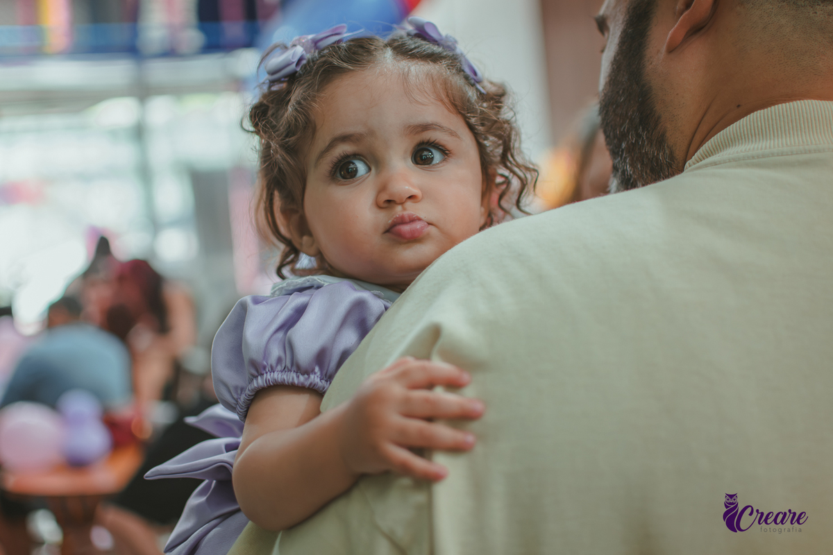 Uma bebê olhando por cima do ombro de um homem, durante festa de aniversário de 1 ano.