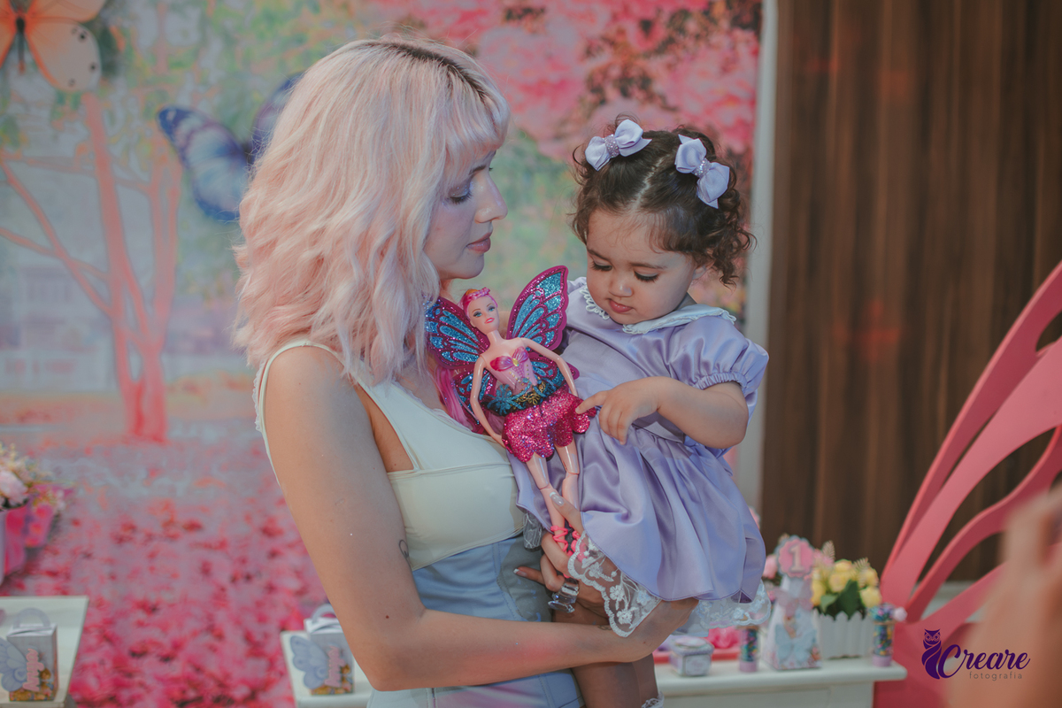 Uma bebê brincando com sua tia e uma barbie borboleta, durante sua festa de anievrsário de 1 ano.