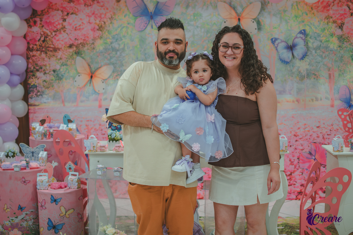Uma casal com sua bebê no colo em frente uma decoração com tema borboletas, durante festa de aniversário de 1 ano.