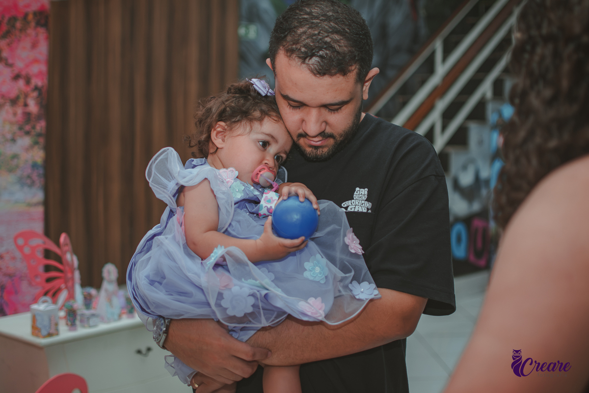 Uma homem aconchegando uma bebê, que está quase dormindo, durante festa de aniversário de 1 ano.