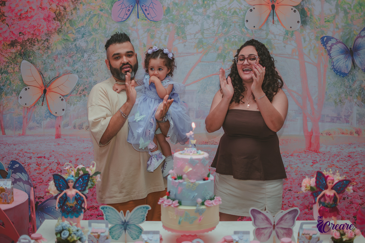 Uma mulher e um homem com bebê no colo, durante hora do parabéns em aniversário de 1 ano.