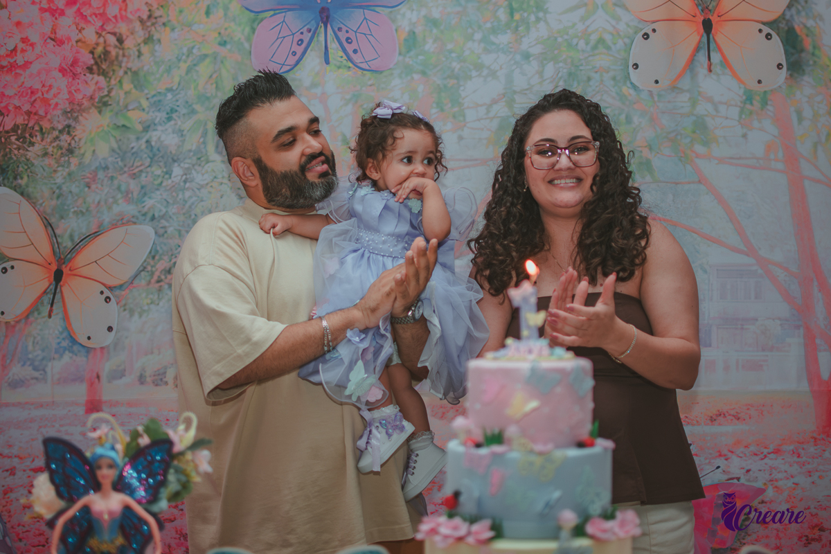 Uma mulher e um homem com bebê no colo, durante hora do parabéns em aniversário de 1 ano.