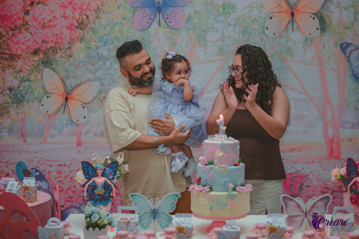 Uma mulher e um homem com bebê no colo, durante hora do parabéns em aniversário de 1 ano.