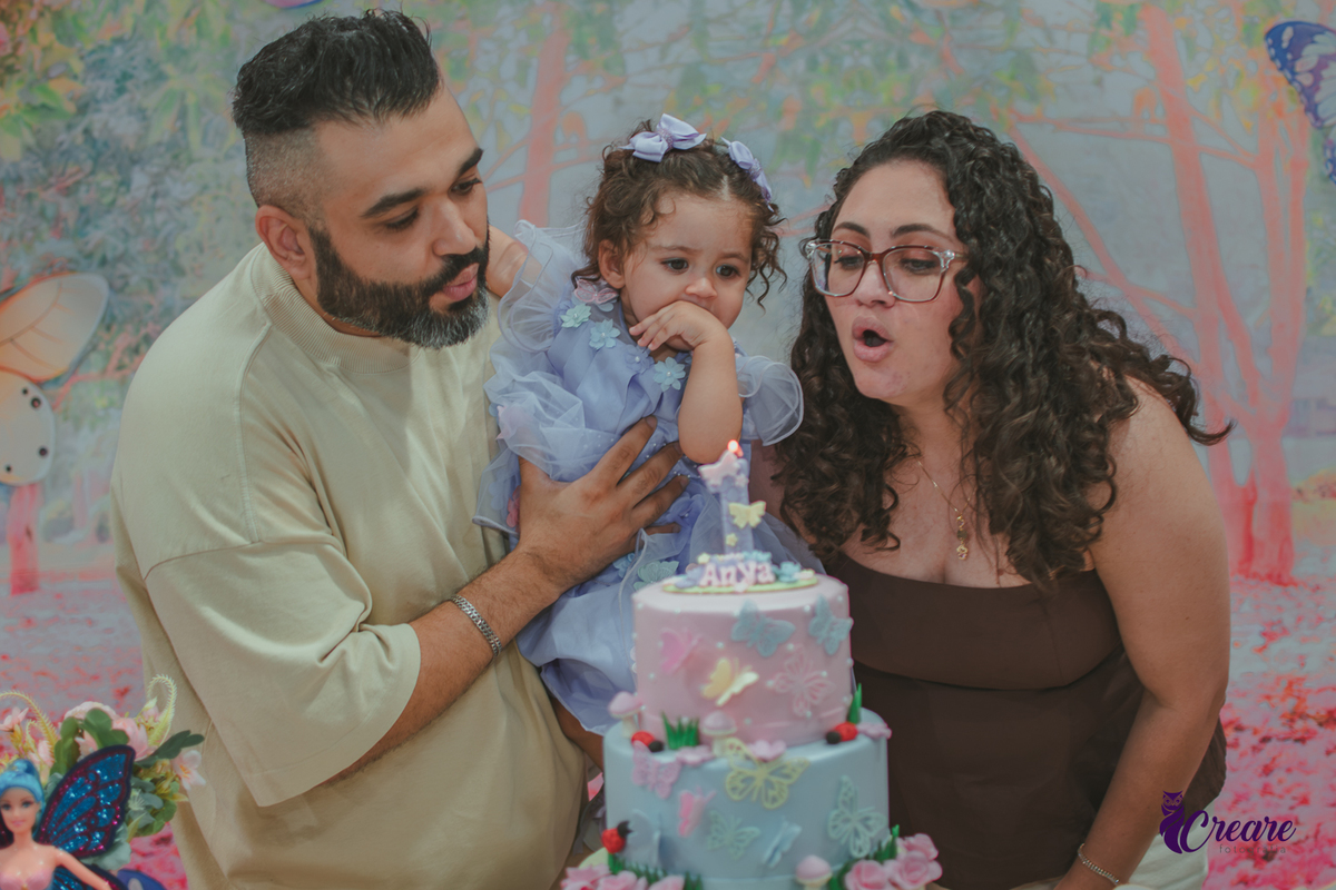 Uma homem com uma bebê no colo e uma mulher soprando uma velinha na hora do parabéns, durante festa de aniversário infantil.