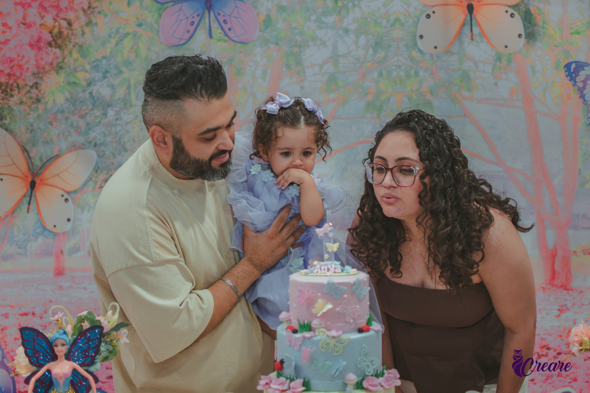 Uma homem com uma bebê no colo e uma mulher soprando uma velinha na hora do parabéns, durante festa de aniversário infantil.