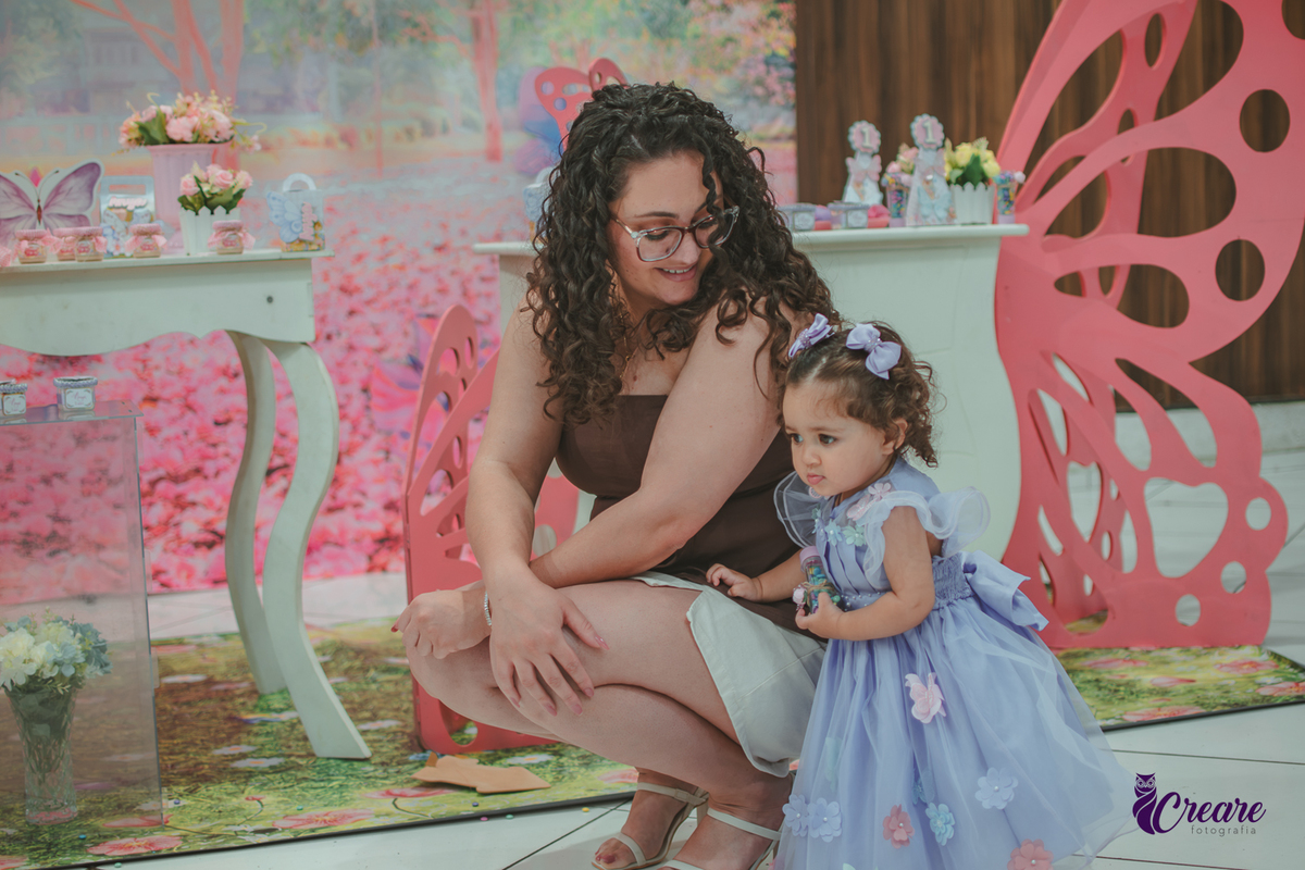 Uma mulher com sua filha ao lado em frente uma decoração de aniversário com tema borboleta, durante festa de aniversário infantil.