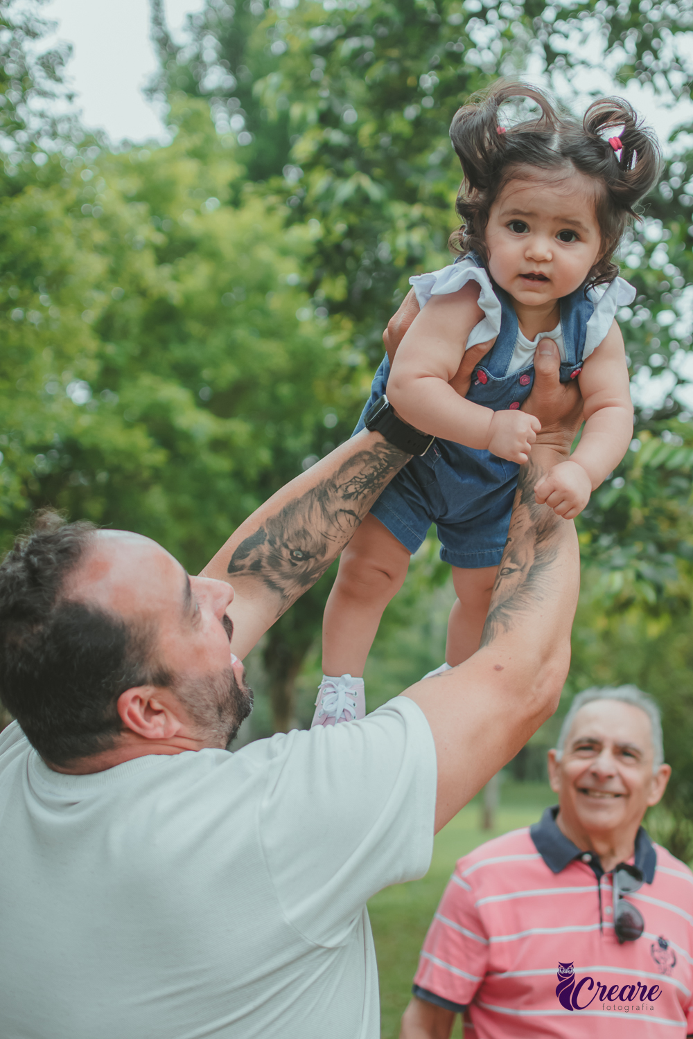 Uma homem brincando de aviãozinho com uma bebê de 1 ano, que está usando uma jardineira azul, durante sua festa de aniversário no Parque Central de Santo André.
