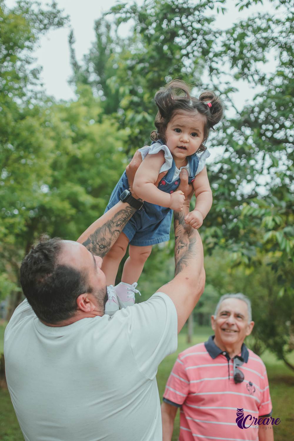 Uma homem brincando de aviãozinho com uma bebê de 1 ano, que está usando uma jardineira azul, durante sua festa de aniversário no Parque Central de Santo André.