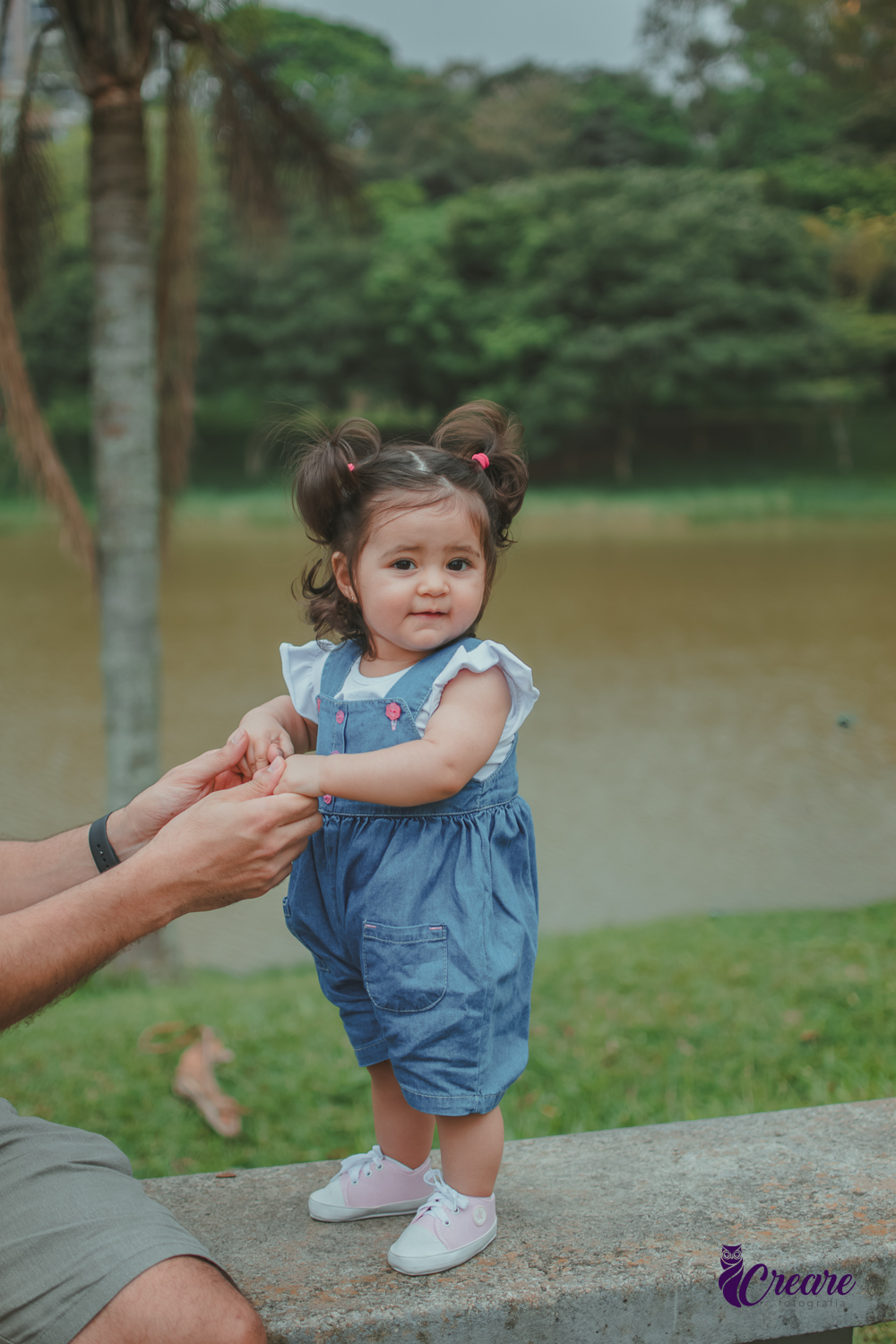 Bebê sorrindo em pé em um banco do Parque Central de Santo André, com um lago ao fundo, durante a comemoração de seu aniversário de 1 ano ao ar livre.