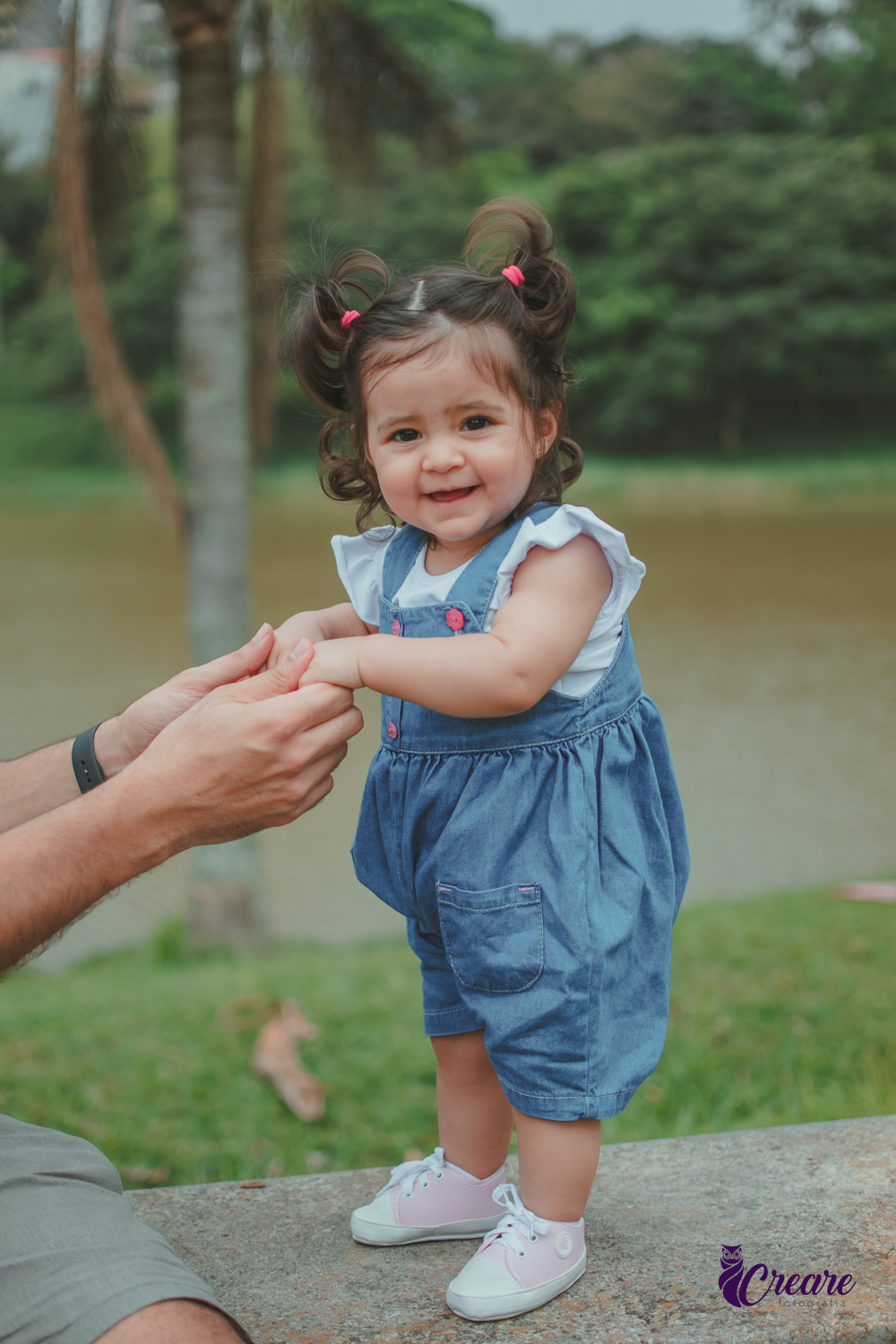 Bebê sorrindo em pé em um banco do Parque Central de Santo André, com um lago ao fundo, durante a comemoração de seu aniversário de 1 ano ao ar livre.