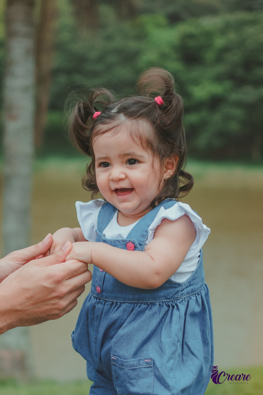 Bebê sorrindo em pé em um banco do Parque Central de Santo André, com um lago ao fundo, durante a comemoração de seu aniversário de 1 ano ao ar livre.