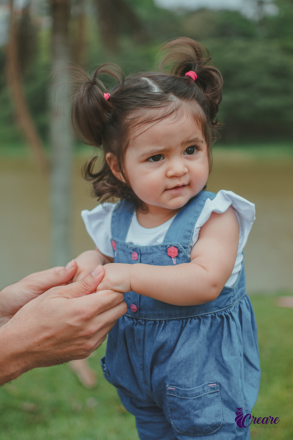 Bebê sorrindo em pé em um banco do Parque Central de Santo André, com um lago ao fundo, durante a comemoração de seu aniversário de 1 ano ao ar livre.