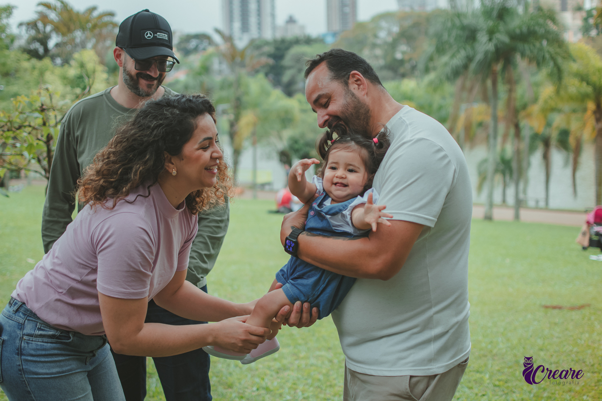Homem segurando uma bebê no colo enquanto uma mulher brinca com ela durante a comemoração de seu primeiro aniversário no Parque Central de Santo André.