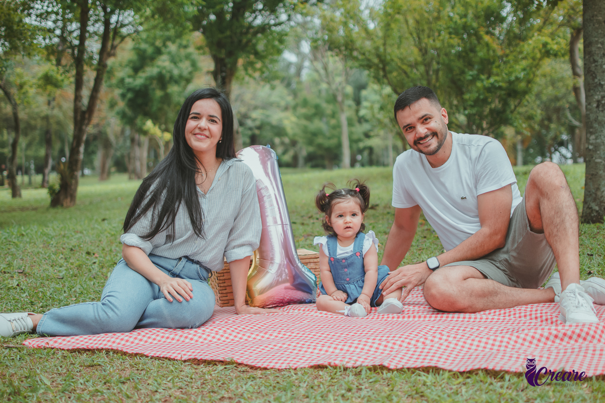 Bebê de jardineira azul, junto com seus pais, sentada sobre uma toalha vermelha de piquenique, ao lado de um balão de número 1 e uma cesta de piquenique, durante a comemoração de seu primeiro aniversário no Parque Central de Santo André.