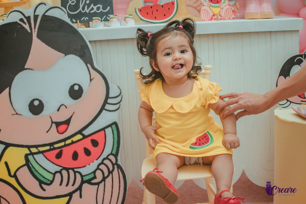 Menina de vestido amarelo em frente à decoração de aniversário de 1 ano com tema Magali, em uma festa infantil.