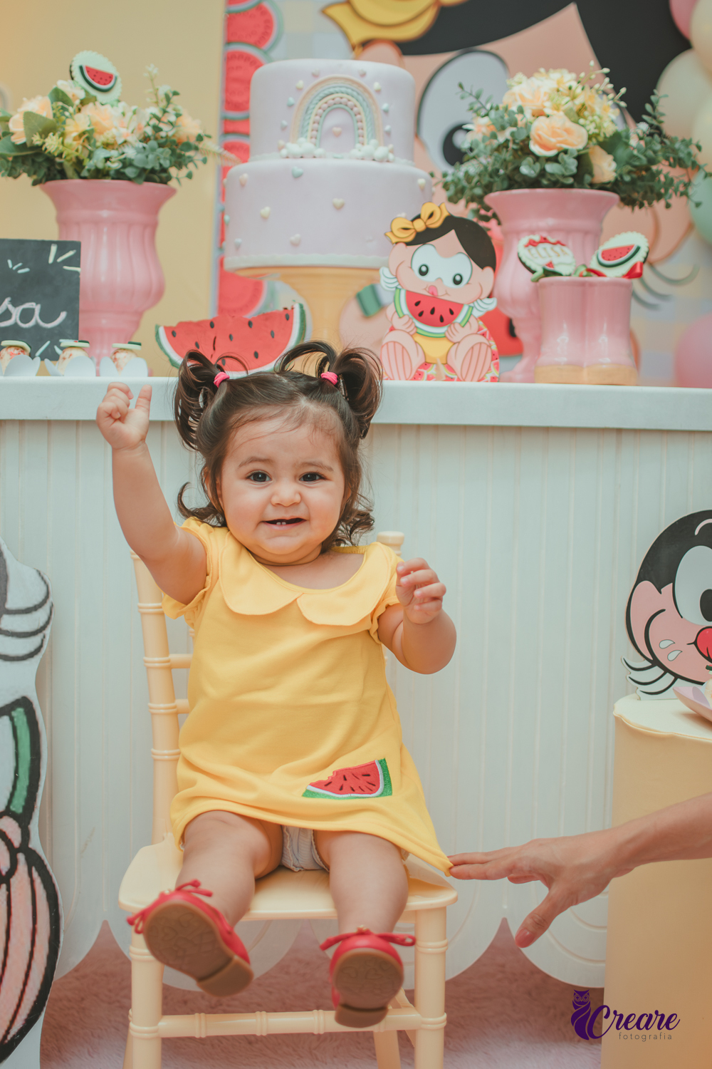 Menina de vestido amarelo em frente à decoração de aniversário de 1 ano com tema Magali, em uma festa infantil.
