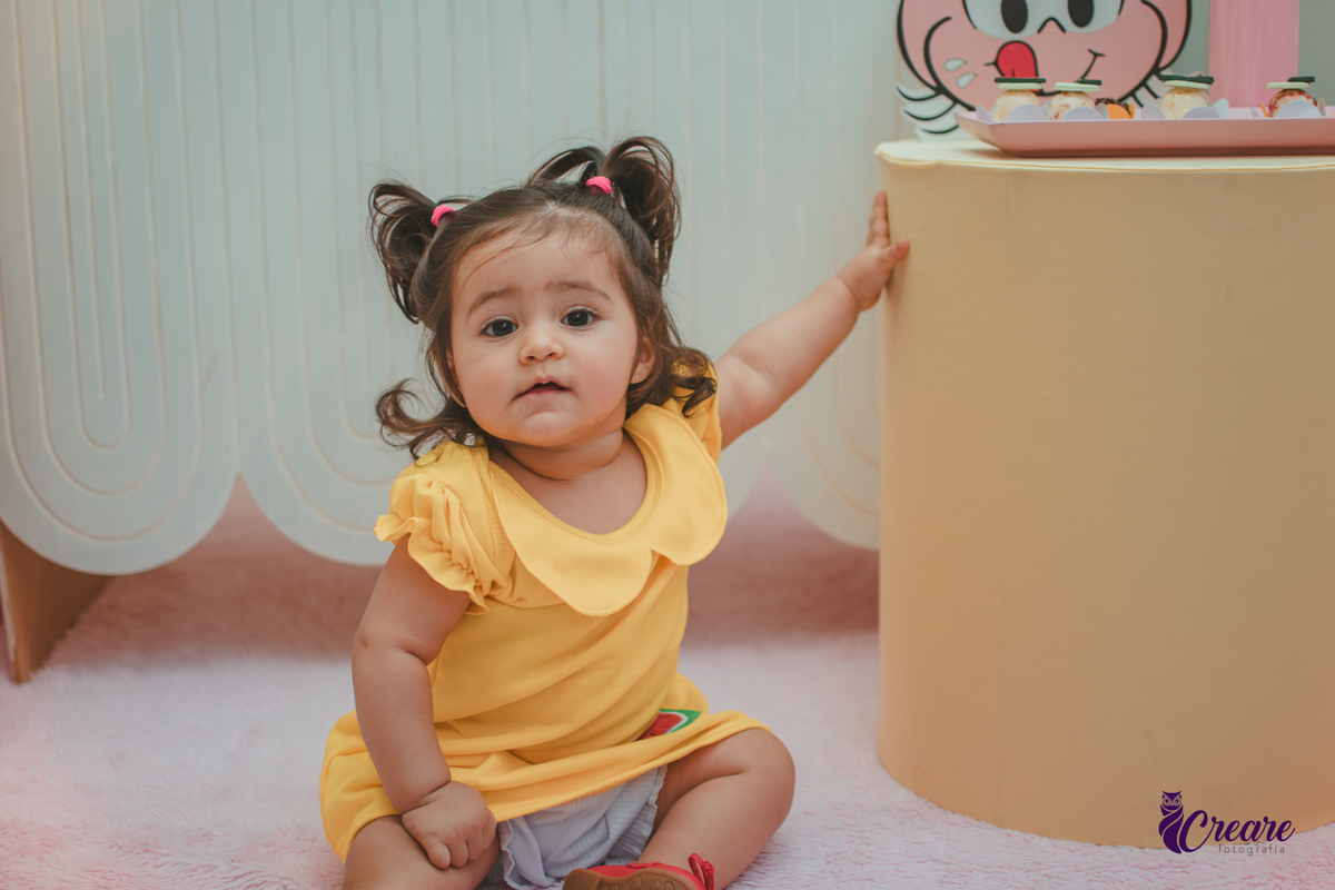 Menina de vestido amarelo em frente à decoração de aniversário de 1 ano com tema Magali, em uma festa infantil.