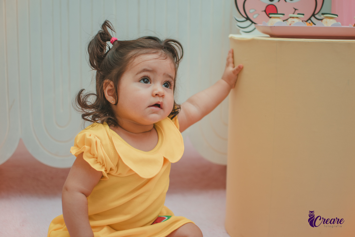 Menina de vestido amarelo em frente à decoração de aniversário de 1 ano com tema Magali, em uma festa infantil.
