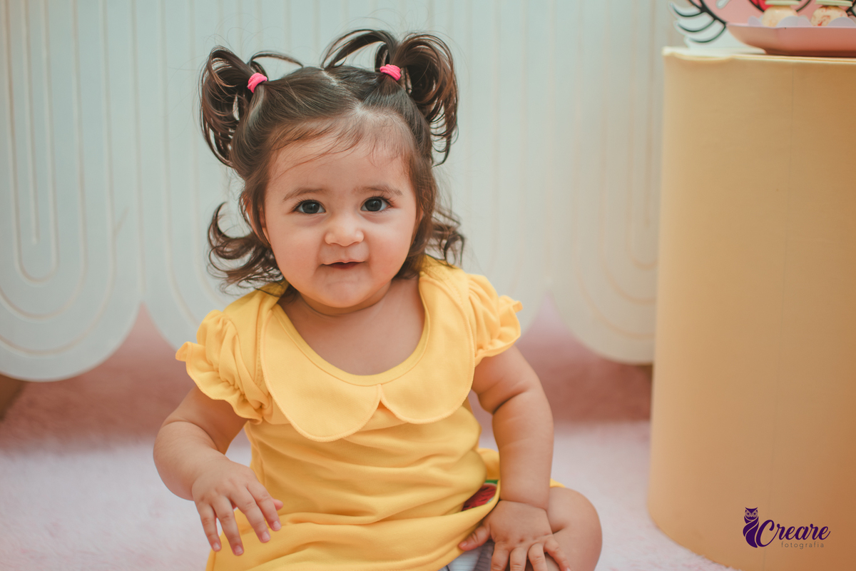 Menina de vestido amarelo em frente à decoração de aniversário de 1 ano com tema Magali, em uma festa infantil.