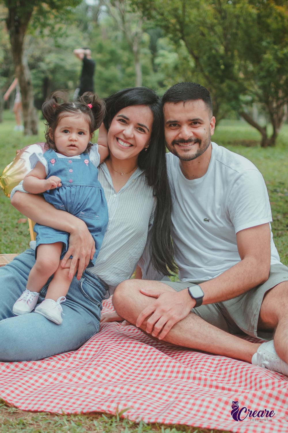 Bebê de jardineira azul, junto com seus pais, sentada sobre uma toalha vermelha de piquenique, ao lado de um balão de número 1 e uma cesta de piquenique, durante a comemoração de seu primeiro aniversário no Parque Central de Santo André.