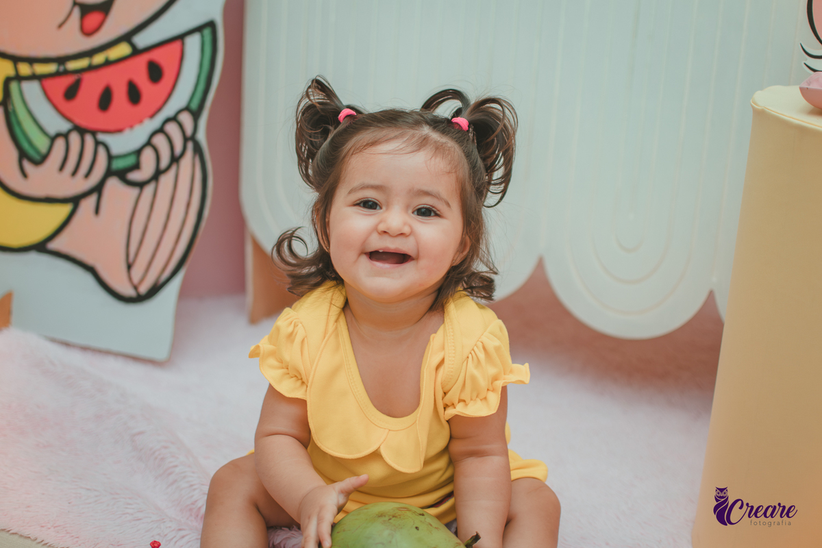 Menina de vestido amarelo em frente à decoração de aniversário de 1 ano com tema Magali, em uma festa infantil.