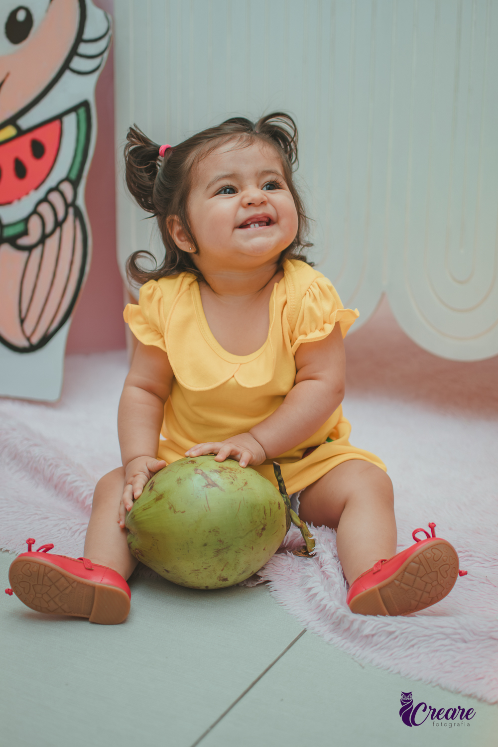 Menina de vestido amarelo, segurando um coco verde, em frente à decoração de aniversário de 1 ano com tema Magali, em uma festa infantil.