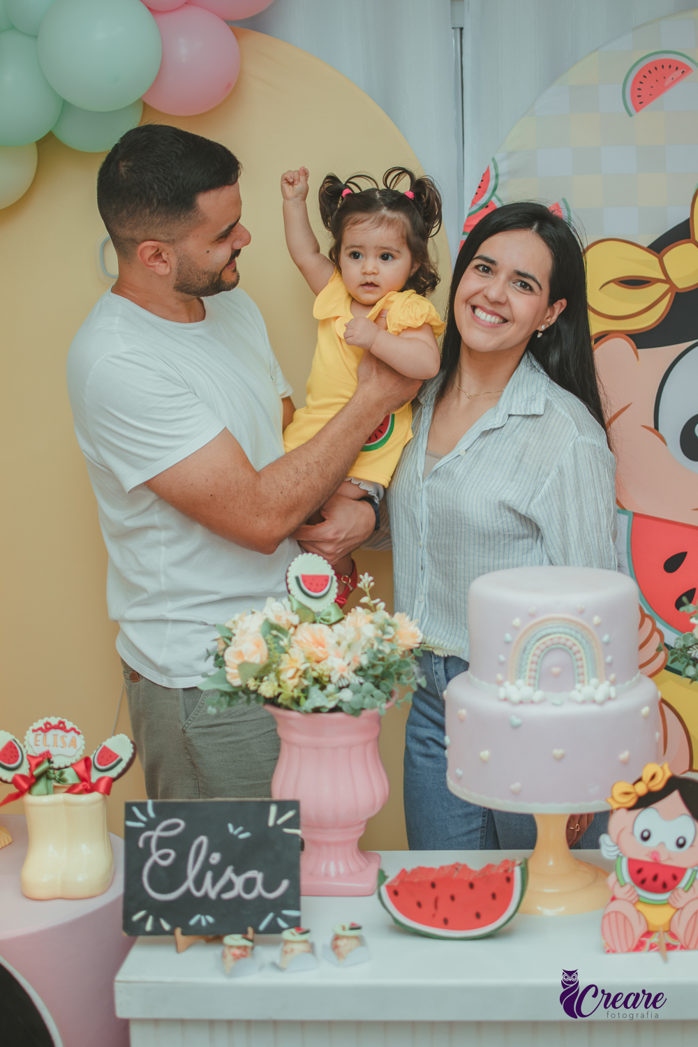 Um casal segurando uma bebê que usa vestido amarelo em frente à decoração de aniversário de 1 ano com tema Magali, em uma festa infantil.