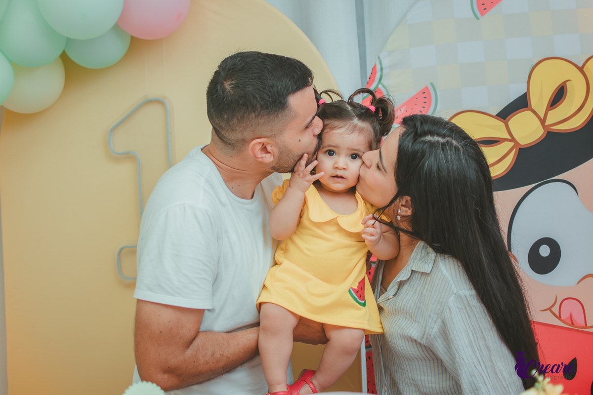 Um casal beijando a bochecha uma bebê que usa vestido amarelo em frente à decoração de aniversário de 1 ano com tema Magali, em uma festa infantil.