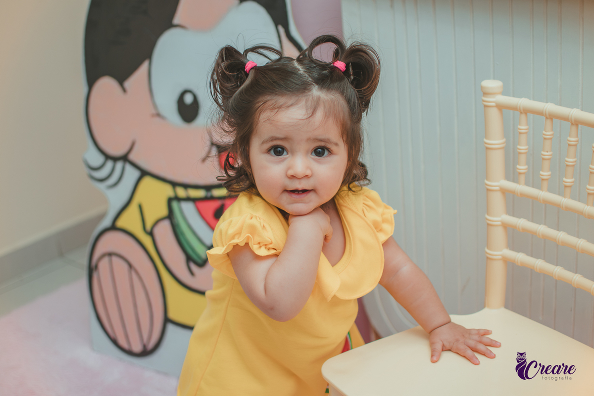 Menina de vestido amarelo em frente à decoração de aniversário de 1 ano com tema Magali, em uma festa infantil.