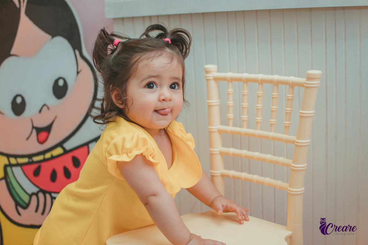 Menina de vestido amarelo em frente à decoração de aniversário de 1 ano com tema Magali, em uma festa infantil.