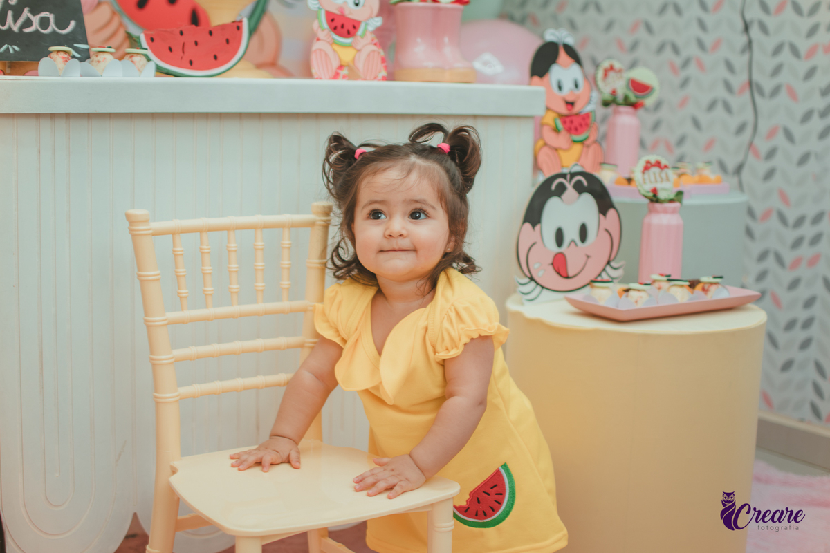 Menina de vestido amarelo em frente à decoração de aniversário de 1 ano com tema Magali, em uma festa infantil.