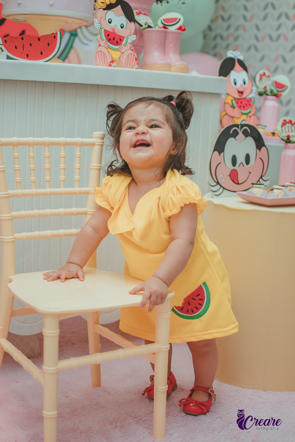Menina de vestido amarelo em frente à decoração de aniversário de 1 ano com tema Magali, em uma festa infantil.