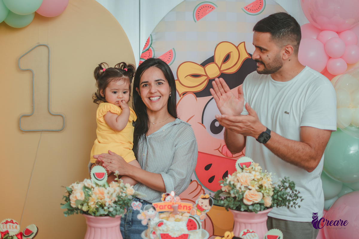 Um casal segurando uma bebê que usa vestido amarelo em frente à decoração de aniversário de 1 ano com tema Magali, durante hora do parabéns.