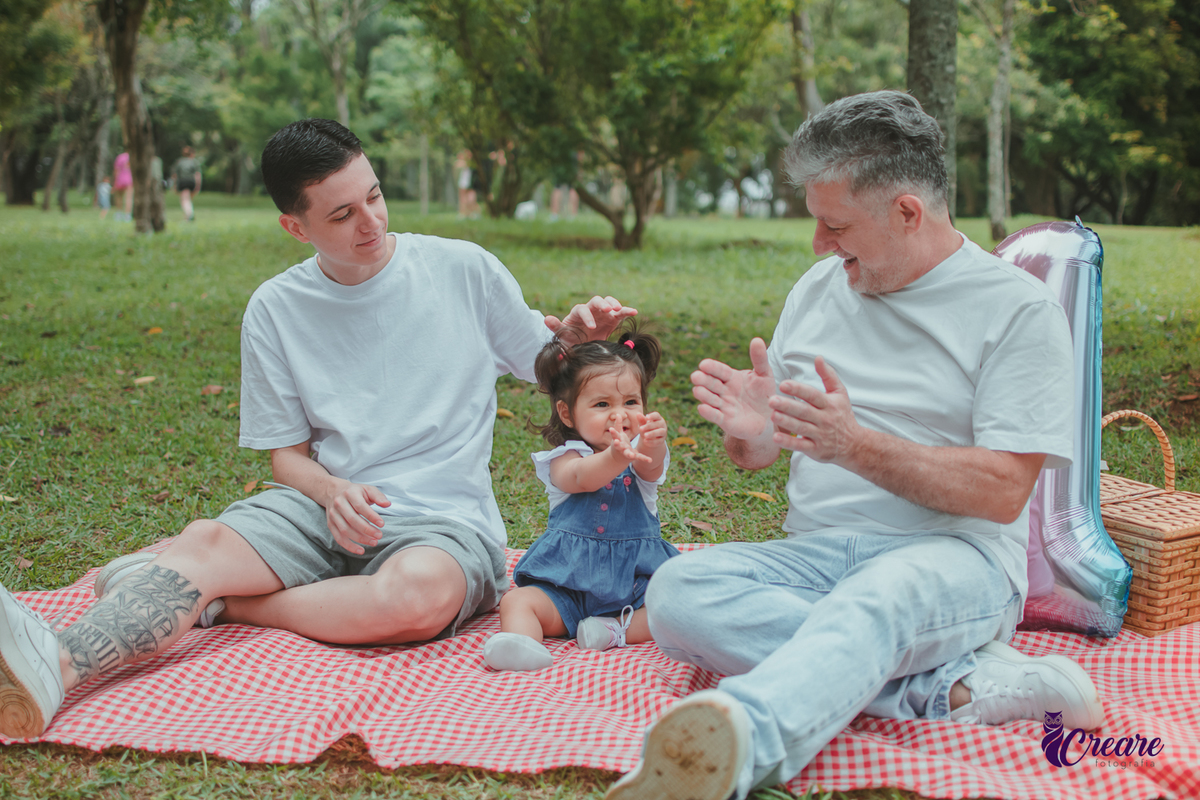 Bebê de jardineira azul, junto com dois homems, sentada sobre uma toalha vermelha de piquenique, ao lado de um balão de número 1 e uma cesta de piquenique, durante a comemoração de seu primeiro aniversário no Parque Central de Santo André.
