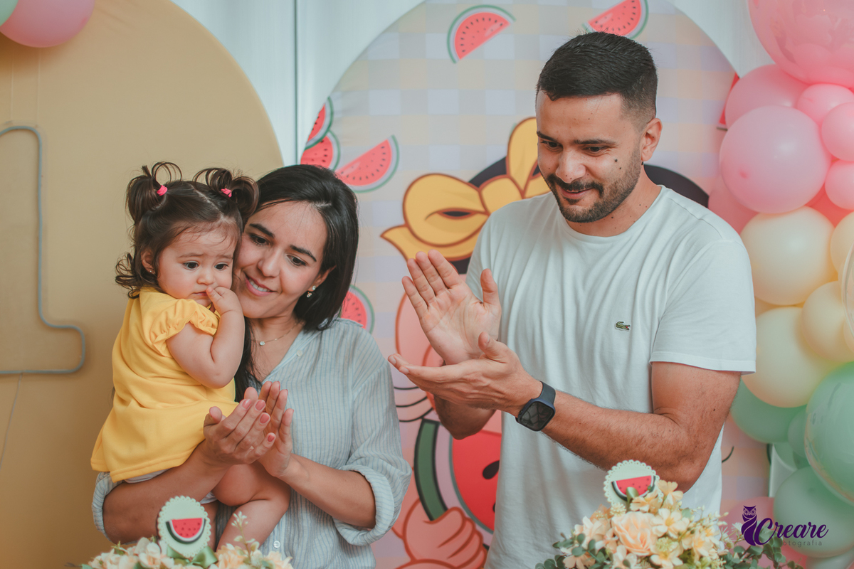 Um casal segurando uma bebê que usa vestido amarelo em frente à decoração de aniversário de 1 ano com tema Magali, durante hora do parabéns.