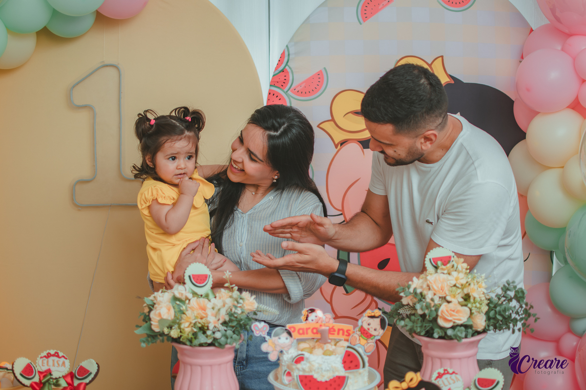 Um casal segurando uma bebê que usa vestido amarelo em frente à decoração de aniversário de 1 ano com tema Magali, durante hora do parabéns.