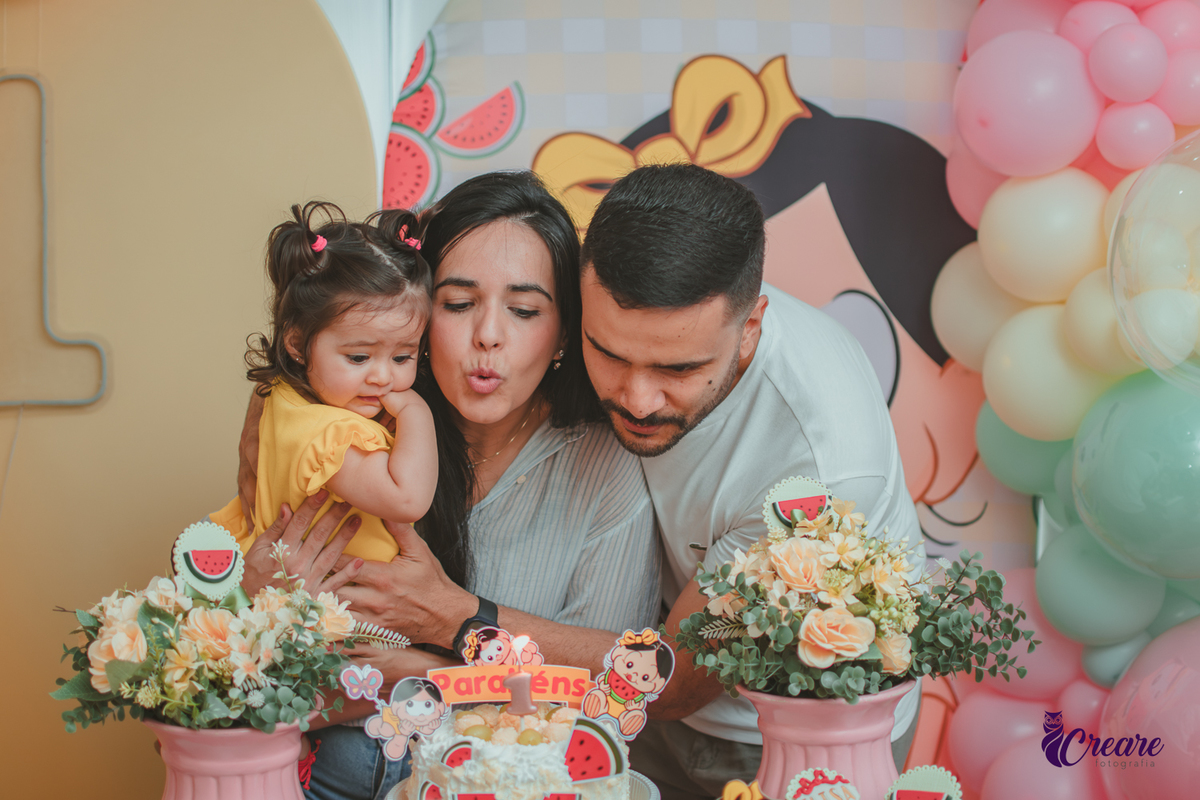 Um casal segurando uma bebê que usa vestido amarelo em frente à decoração de aniversário de 1 ano com tema Magali, durante hora do parabéns.