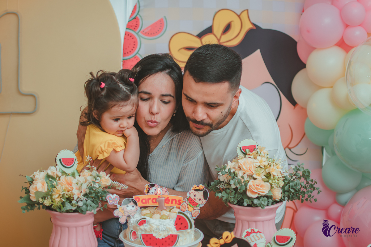 Um casal soprando a vela com uma bebê que usa vestido amarelo em frente à decoração de aniversário de 1 ano com tema Magali, durante hora do parabéns.