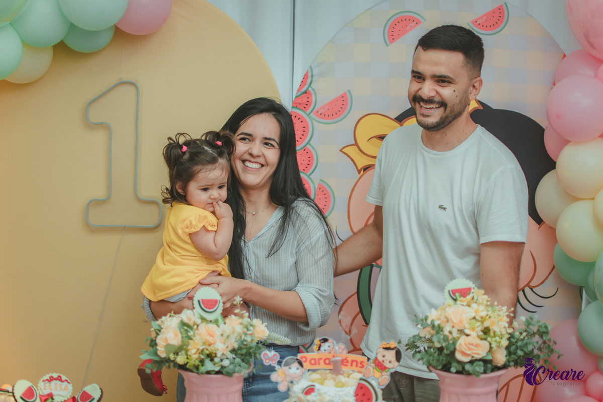 Um casal soprando a vela com uma bebê que usa vestido amarelo em frente à decoração de aniversário de 1 ano com tema Magali, durante hora do parabéns.