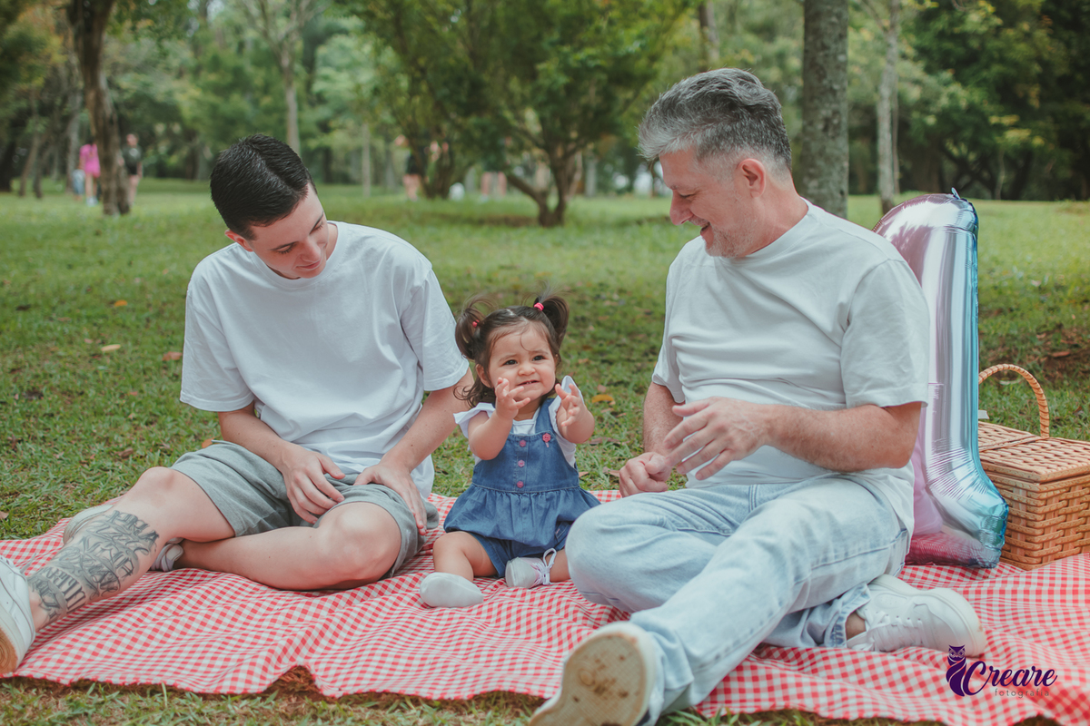 Bebê de jardineira azul, junto com dois homems, sentada sobre uma toalha vermelha de piquenique, ao lado de um balão de número 1 e uma cesta de piquenique, durante a comemoração de seu primeiro aniversário no Parque Central de Santo André.