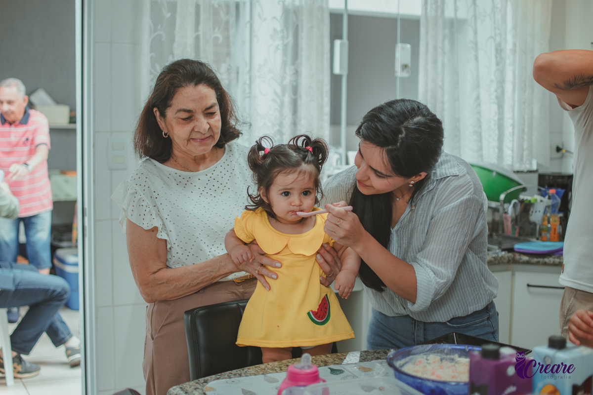 Duas mulheres, dando comida para uma bebê, durante festa de aniversário em casa.