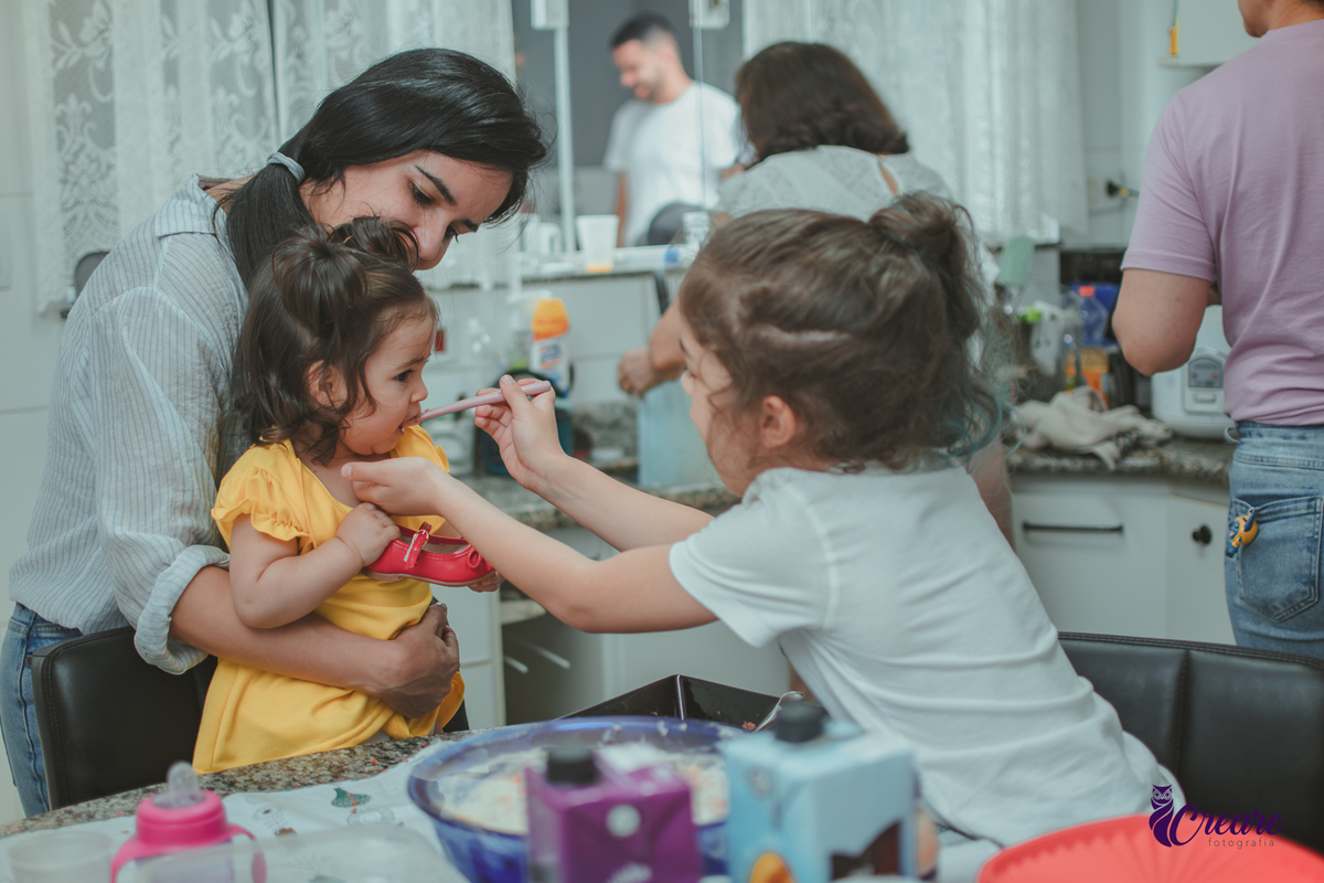 uma mulhere e uma crinaça, dando comida para uma bebê, durante festa de aniversário em casa.
