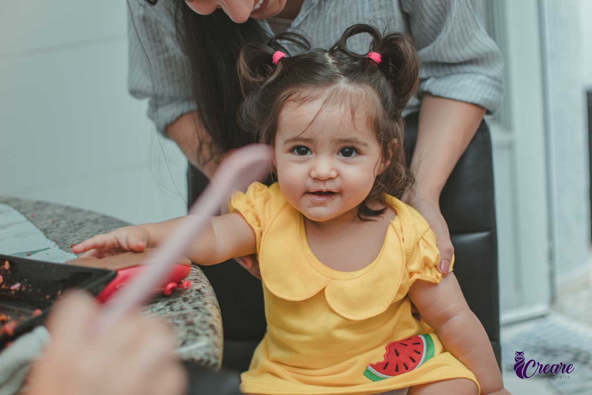 Uma bebê sorrindo enquanto come, durante festa de aniversário de 1 ano em casa.