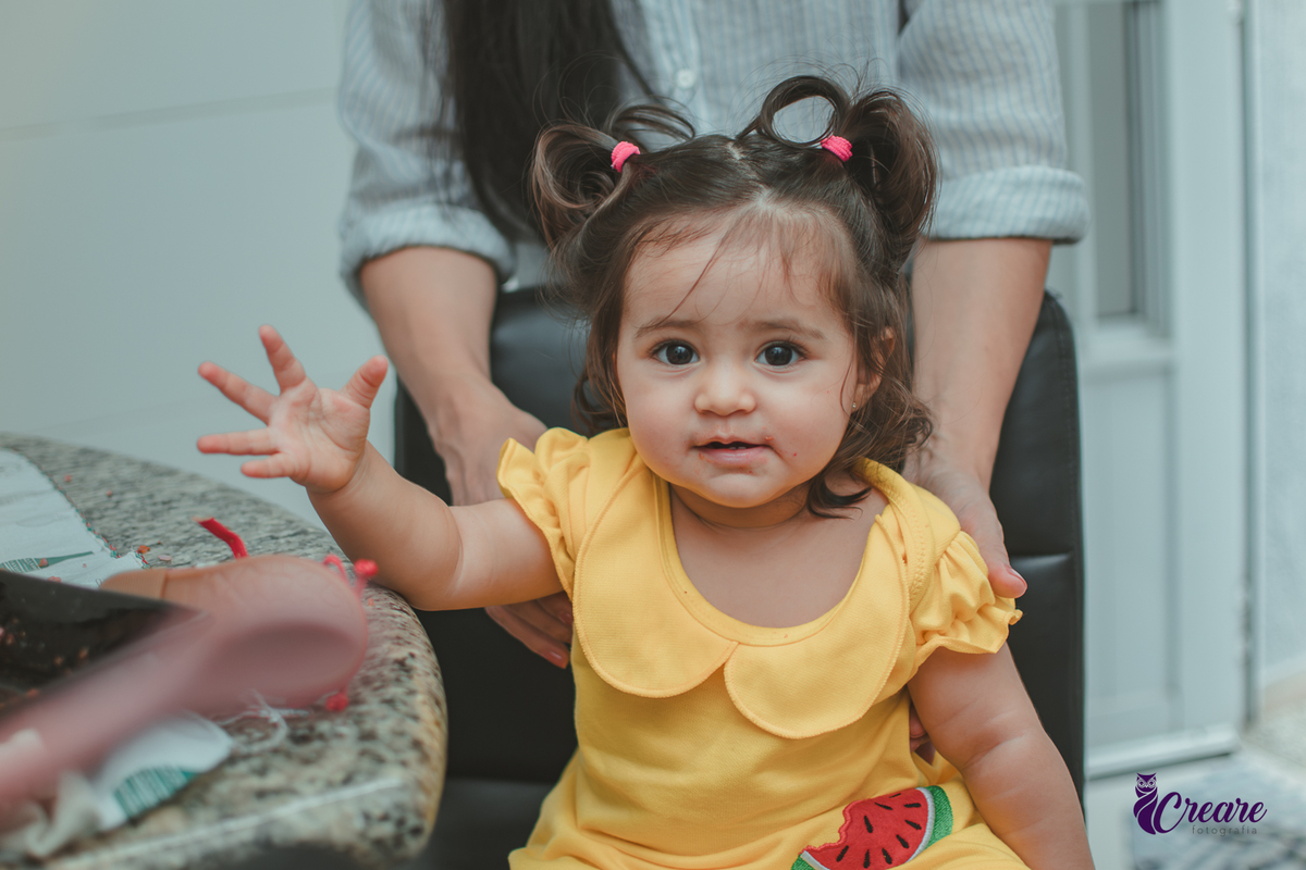 Uma bebê sorrindo enquanto come, durante festa de aniversário de 1 ano em casa.
